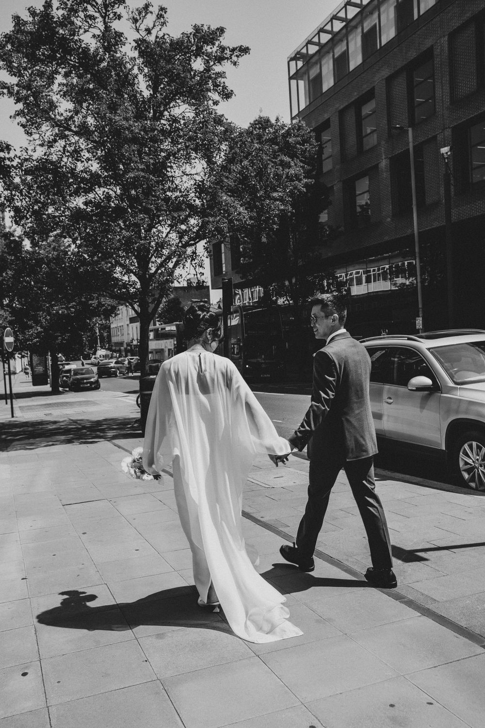 Black and white photo of a bride and groom walking hand in hand in London city sidewalk, wedding dress trailing behind.