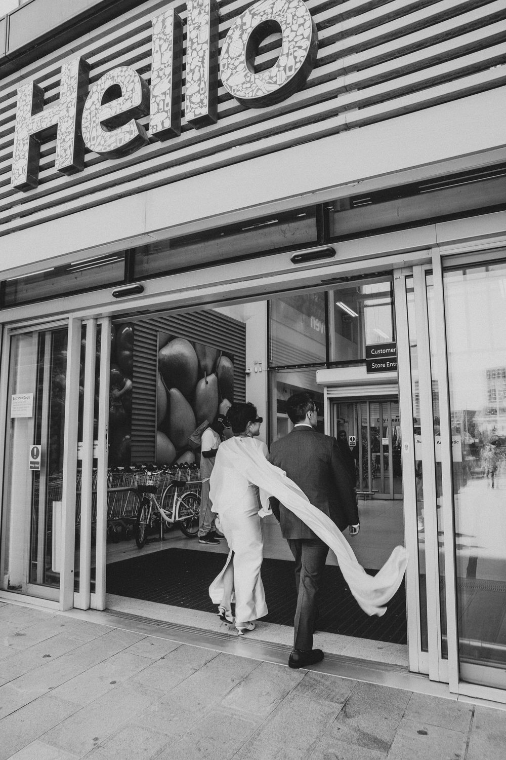 Black and white photo of a bride and groom entering a store with a Hello sign above the sliding doors.