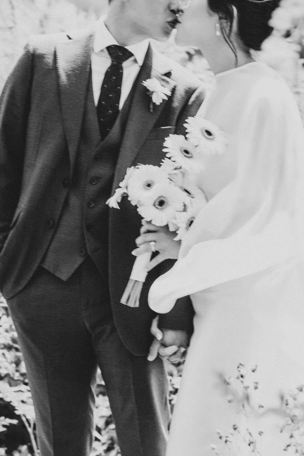 Black and white wedding photo of bride and groom kissing, bride holding gerbera daisy bouquet, groom in three-piece suit.