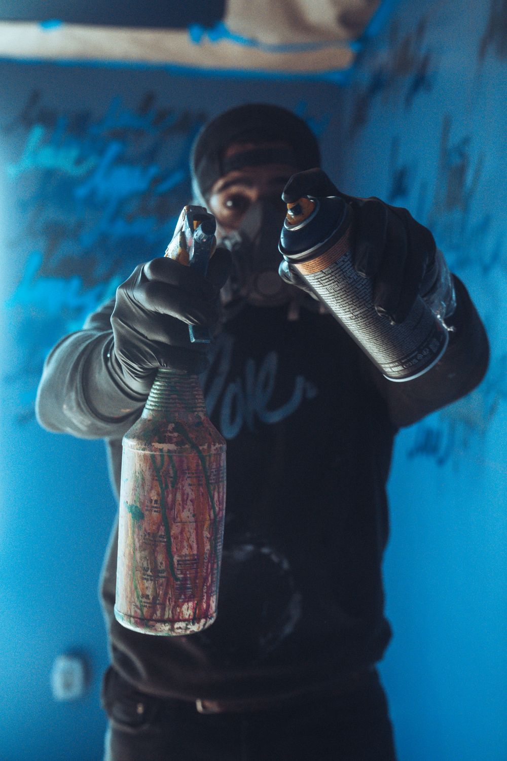 Graffiti artist holding two spray paint cans in a blue-lit room with graffiti-covered walls.