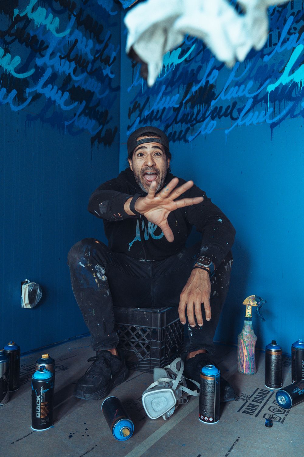 A graffiti artist sits surrounded by spray paint cans against a blue wall covered in street art lettering.