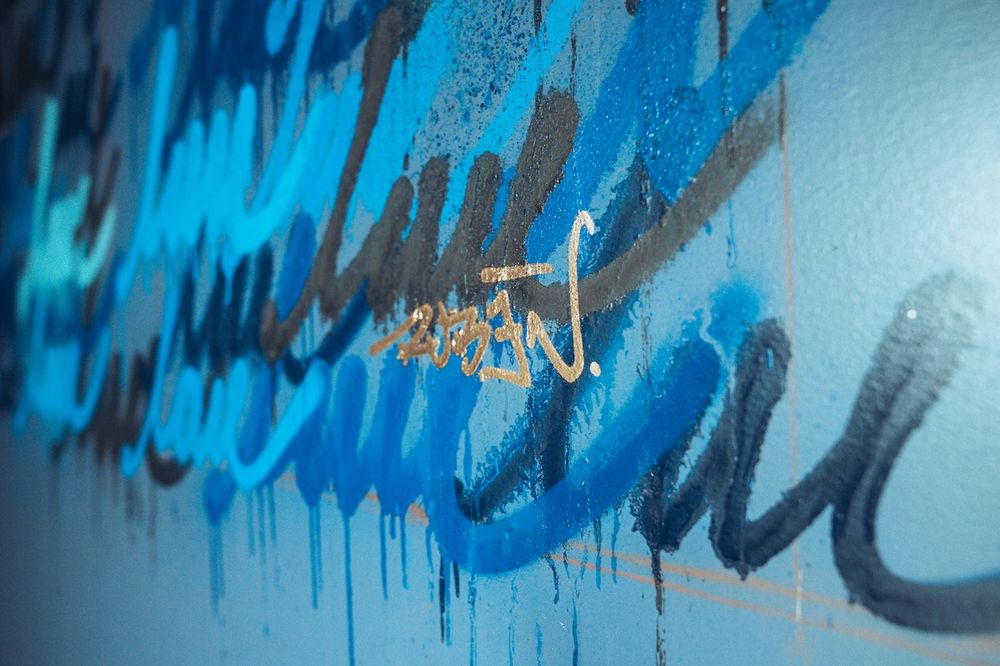 Blue graffiti art with dripping paint on a light blue wall, featuring bold cursive brushstrokes in shades of blue.