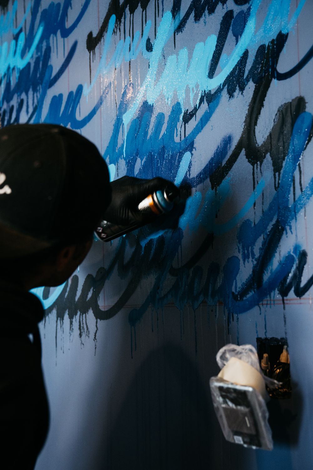 Artist in black cap spray painting blue graffiti calligraphy on a dark wall indoors.