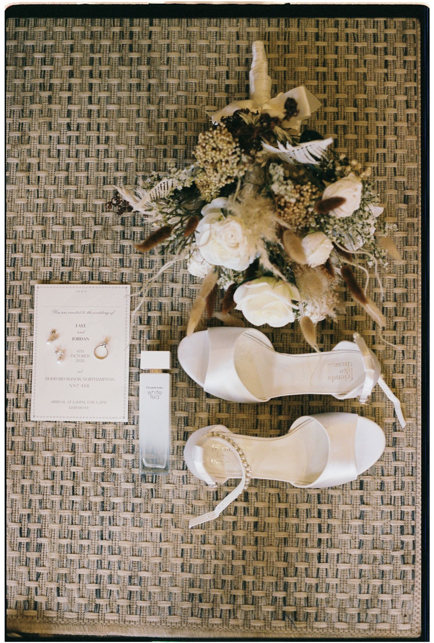 Elegant wedding flat lay featuring white bridal heels, floral bouquet, perfume, and invitation on woven surface.