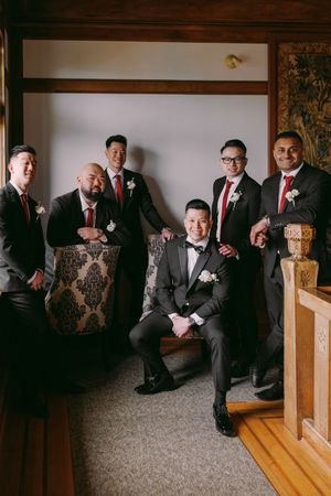 Groom in gray tuxedo poses with five groomsmen in dark suits and red ties in an elegant wedding venue.
