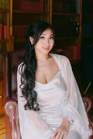 Beautiful Asian bride in white satin gown and lace wrap sits in leather chair in elegant library setting.