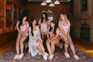 Bride in white robe with bridesmaids in matching pink satin pajamas sitting together in an elegant wood-paneled room.