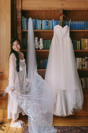 Bride in white robe holds lace veil beside hanging wedding gown and heels in a wood-paneled library.