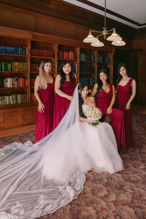 Bride in white gown with veil sits holding bouquet, surrounded by four bridesmaids in red dresses in an elegant library.