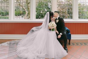 A bride and groom share a romantic kiss in a sunlit venue, her elegant white gown and veil flowing gracefully around them.