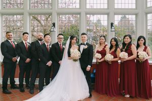 Wedding party in burgundy and black attire poses indoors, bride in white gown holding bouquet of white roses.