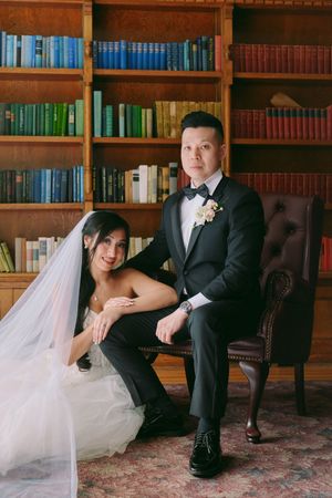 A bride and groom pose elegantly in a library setting, she in a white gown with veil, he in a black tuxedo.