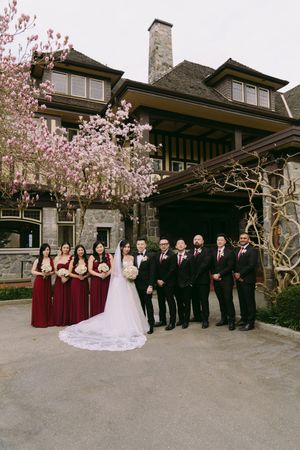 Wedding party poses before a grand estate with blooming cherry blossoms, bridesmaids in burgundy gowns and groomsmen in black suits.