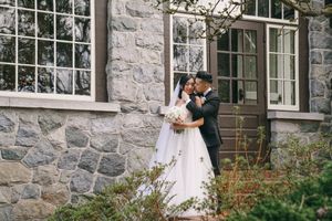 Newlyweds share a romantic embrace outside a stone building surrounded by lush greenery on their wedding day.