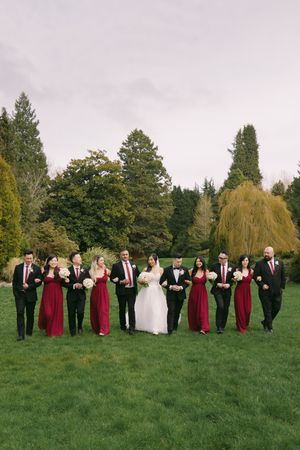 Wedding party walks across green lawn with tall trees, bridesmaids in burgundy dresses, groomsmen in black.
