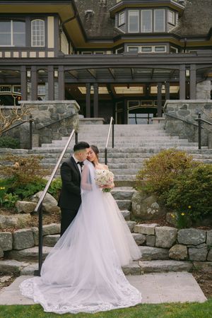 Bride and groom pose on stone steps outside elegant manor, bride in flowing white gown holding bouquet.