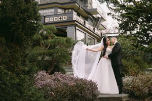 Wedding couple sharing a romantic kiss in a lush garden with veil flowing in the wind.