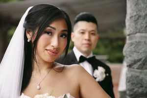 A bride in a white strapless gown and veil smiles softly, with her groom in a black tuxedo standing behind her.