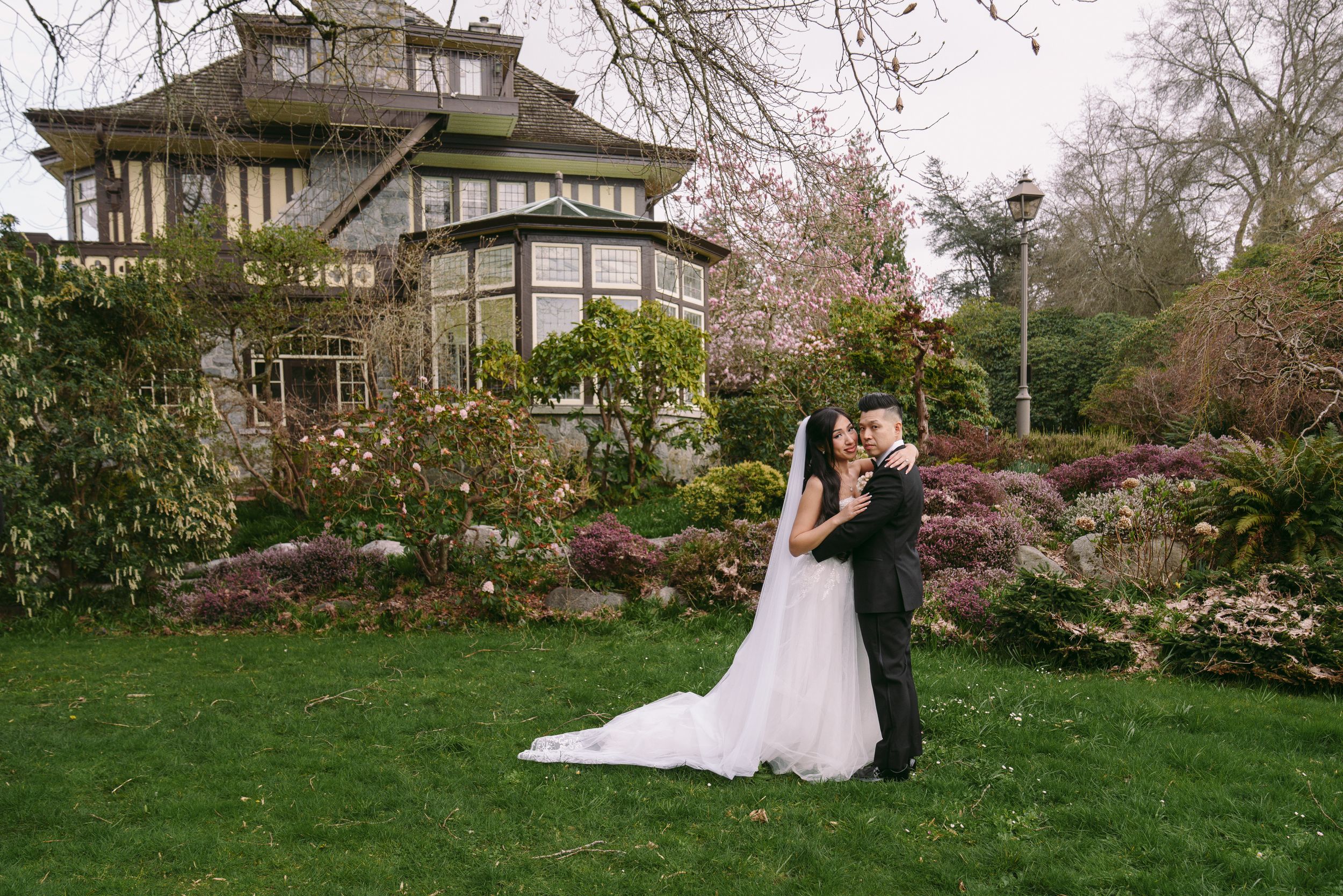 Bride and groom embracing on green lawn before a charming historic manor with spring blooms.