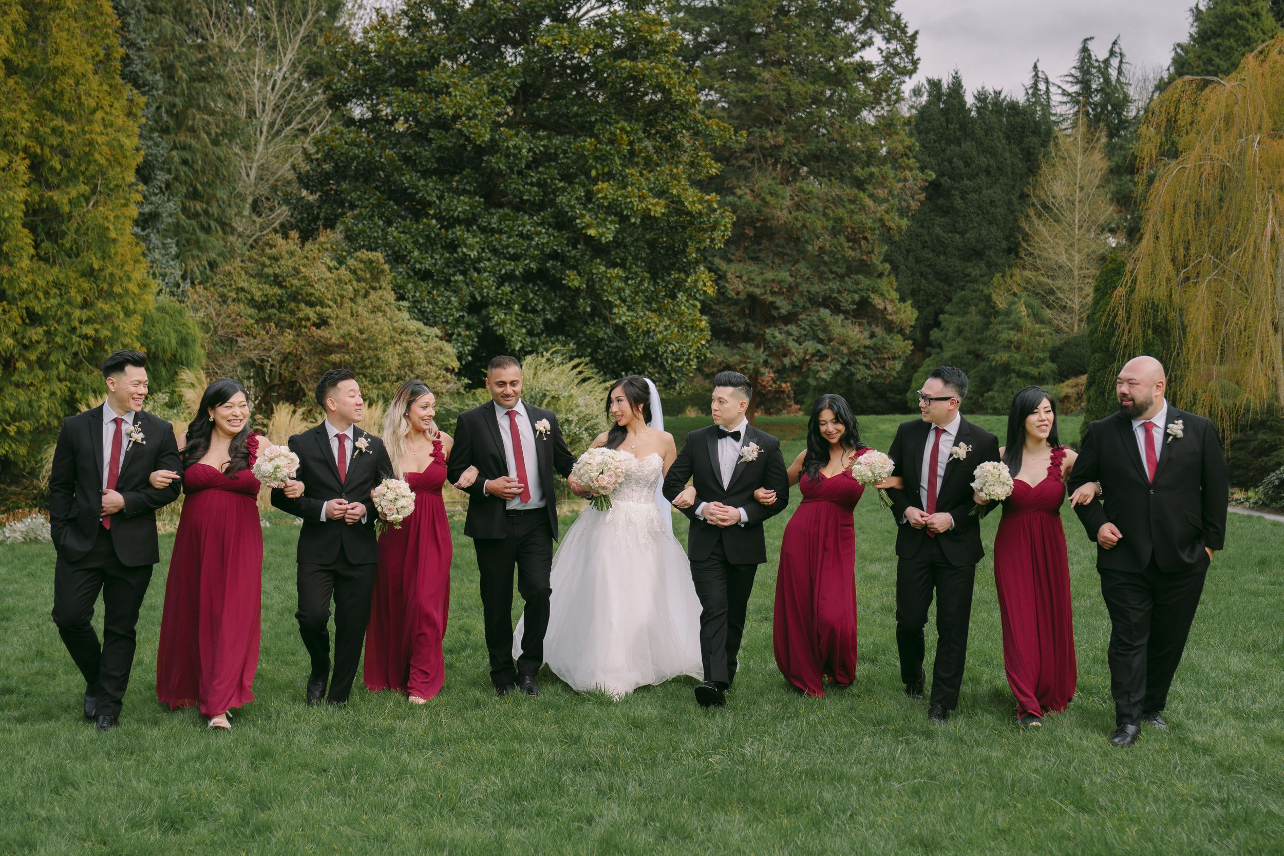 Wedding party in red dresses and black tuxedos walking on green lawn with lush trees in background.