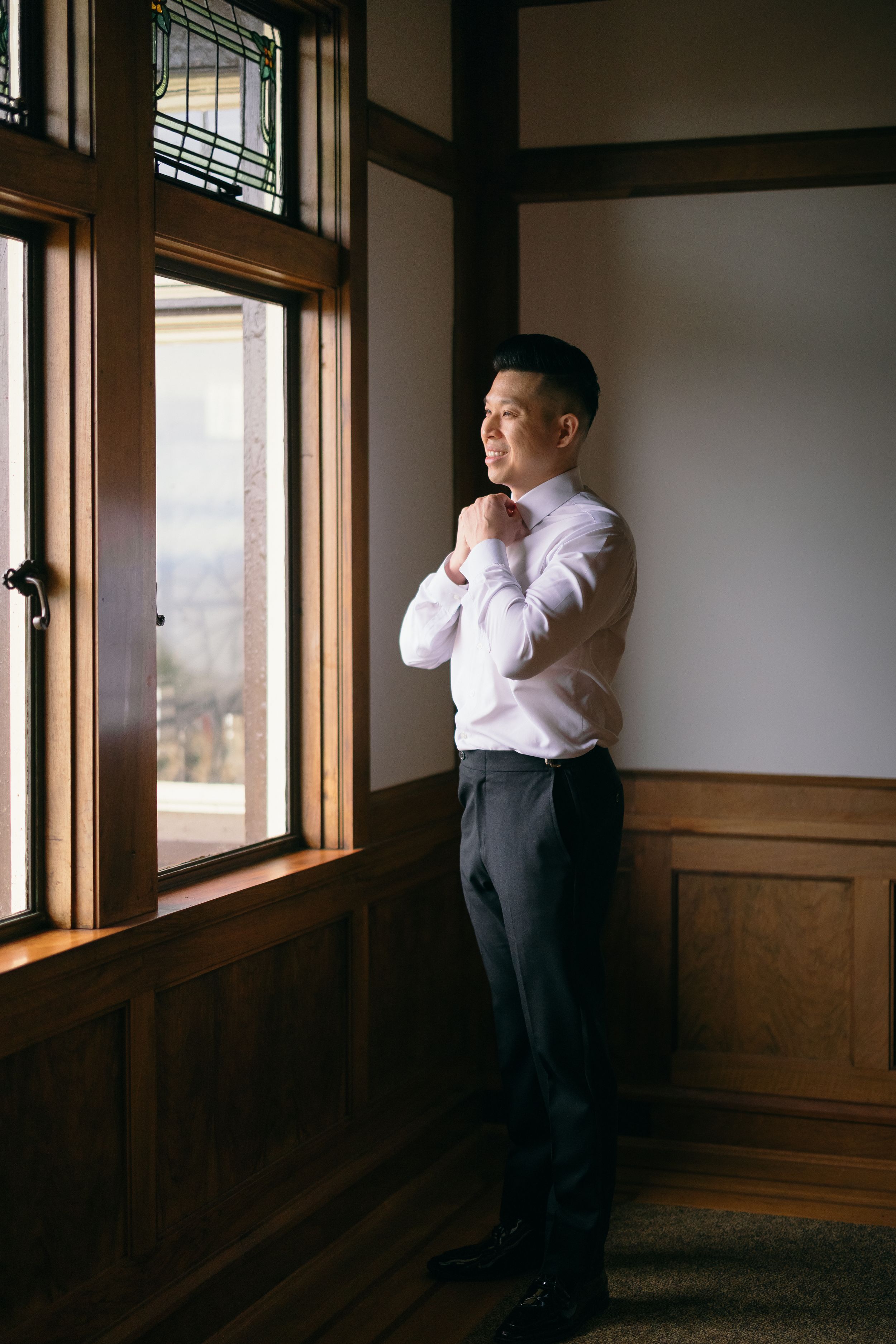 Groom adjusting his collar by a wooden window while getting ready for his wedding day.