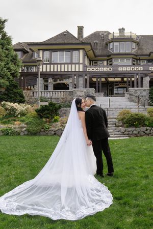Bride and groom sharing a kiss on green lawn before grand Tudor-style estate, long lace cathedral veil trailing behind.