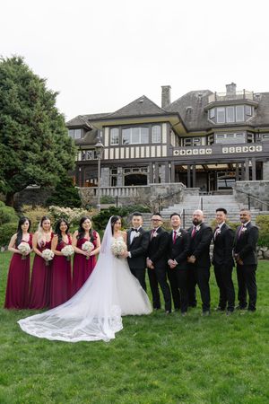 Wedding party in burgundy gowns and black tuxedos pose on lawn before grand estate venue.