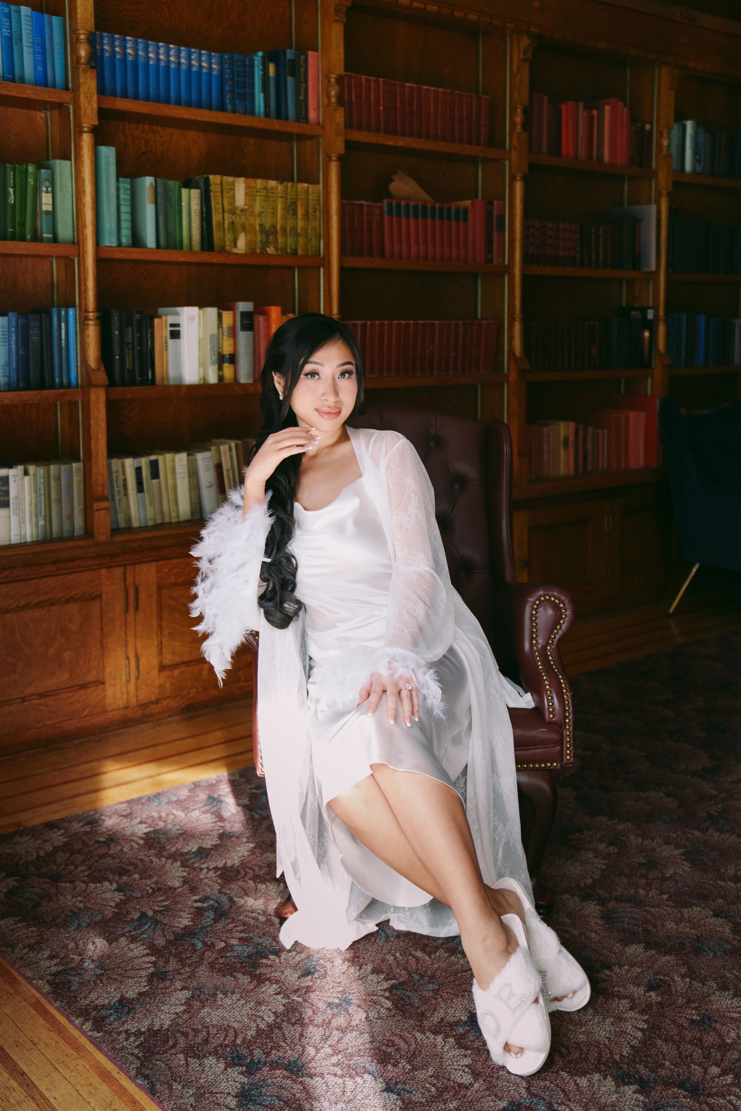 Bride in white robe with feather trim sits elegantly in leather chair before wooden bookshelves in classic library.