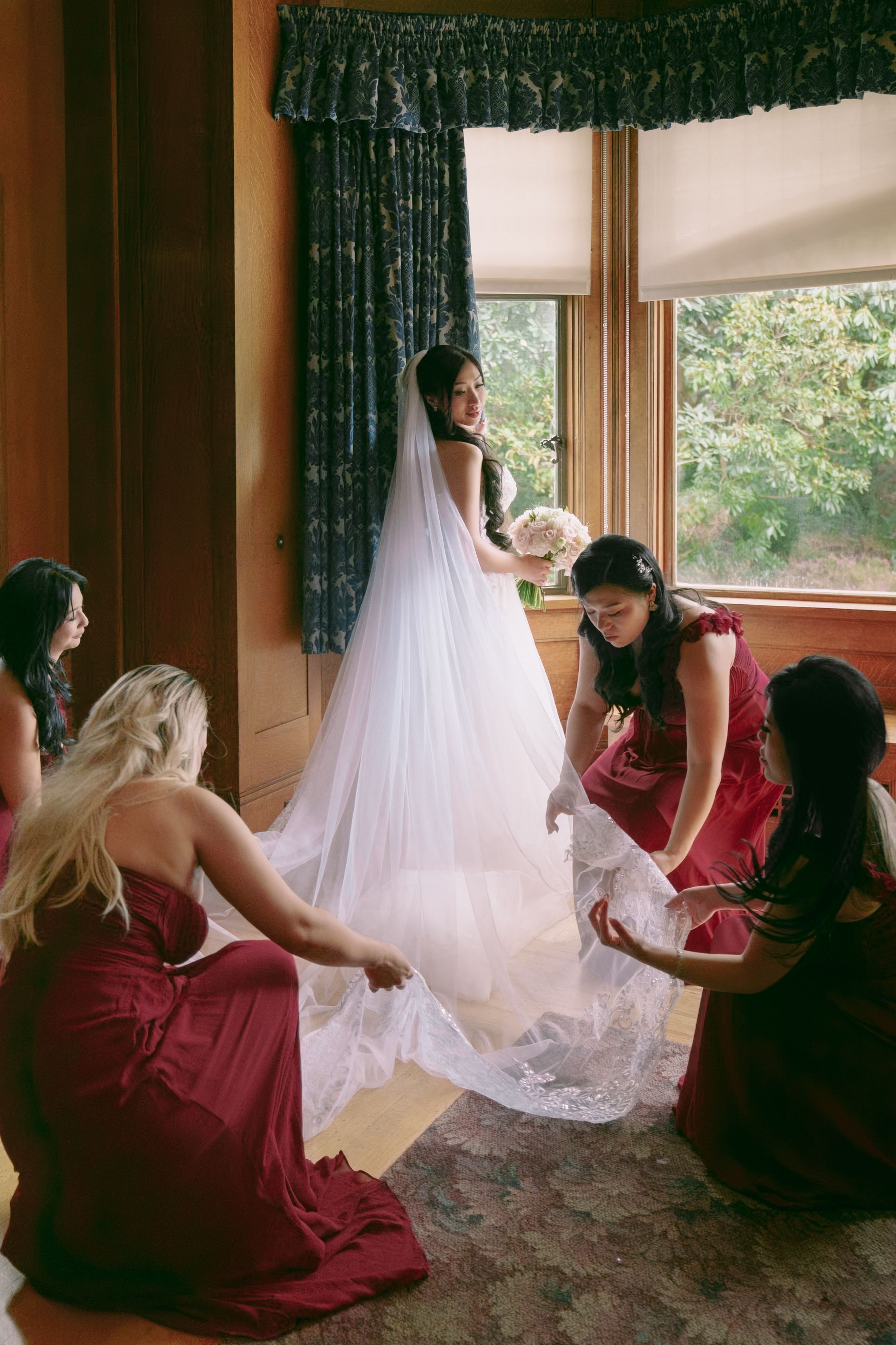 Bride in white gown holds bouquet as bridesmaids in burgundy dresses arrange her veil and train near a sunlit window.
