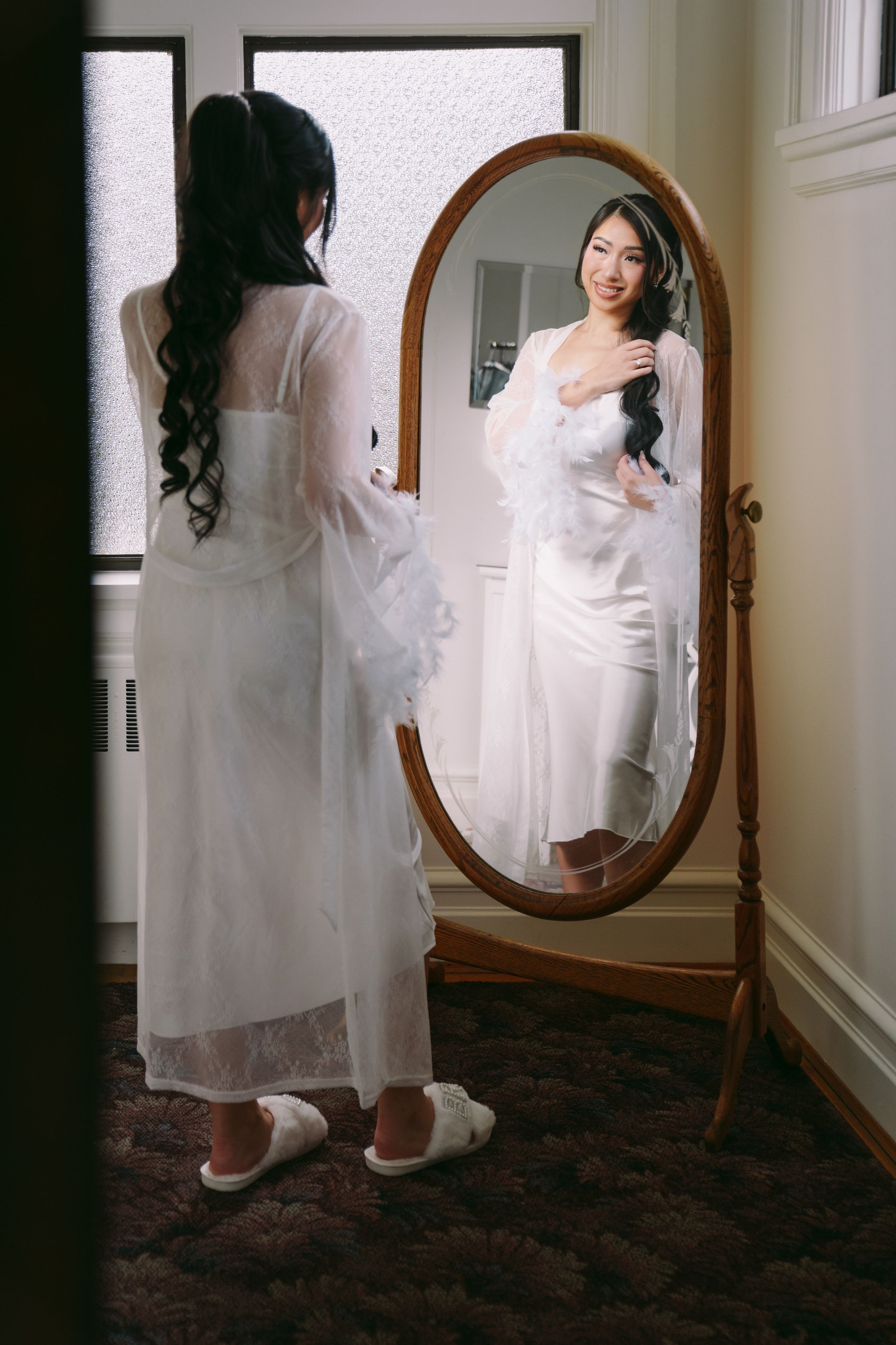 Bride in white lace robe admiring her reflection in an ornate oval floor mirror before her wedding day.