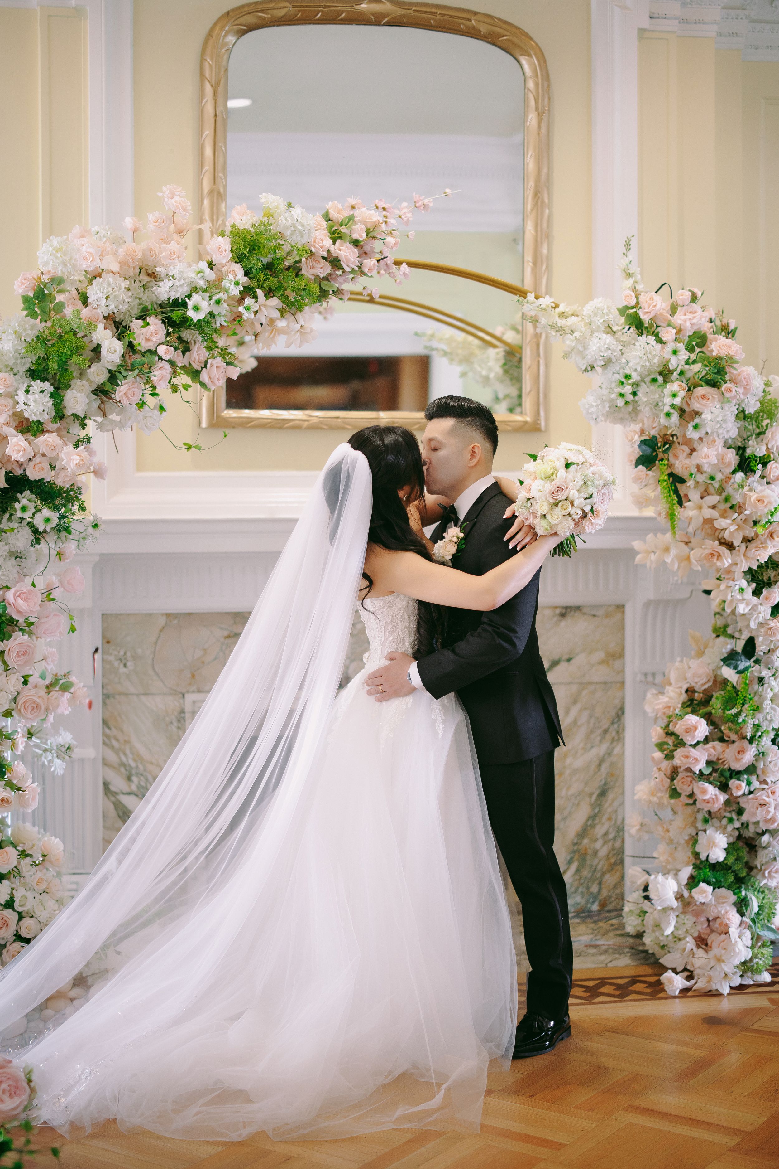 Bride and groom share first kiss under floral arch with pink roses and greenery at elegant indoor wedding ceremony.