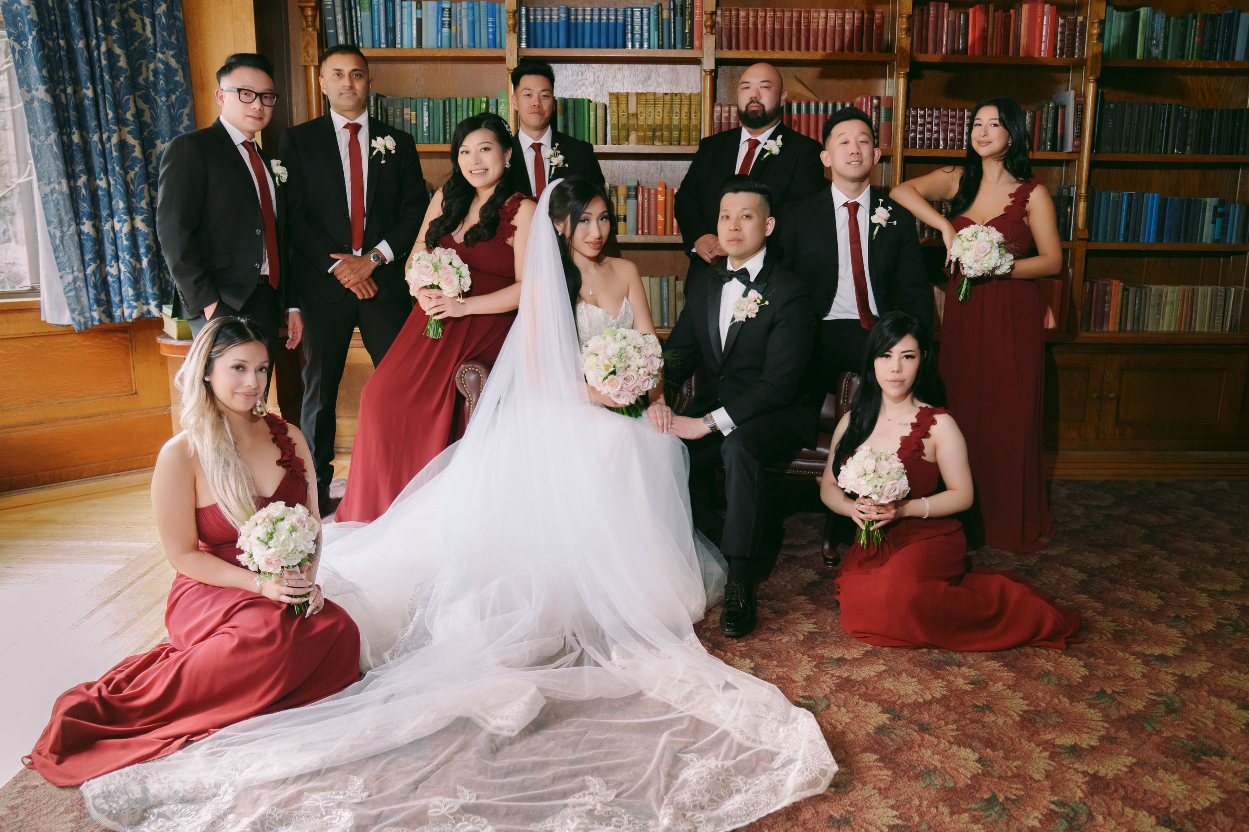Wedding party in burgundy and white poses in a library, bride and groom centered with bridesmaids and groomsmen.