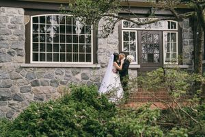 Bride and groom kissing in front of a stone mansion surrounded by lush green gardens on their wedding day.