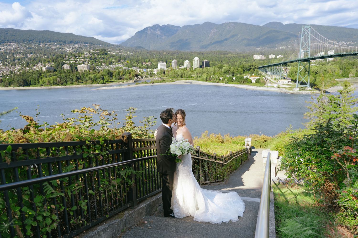 Bride and groom embrace on a scenic overlook with Lions Gate Bridge and Vancouver mountains in the background.