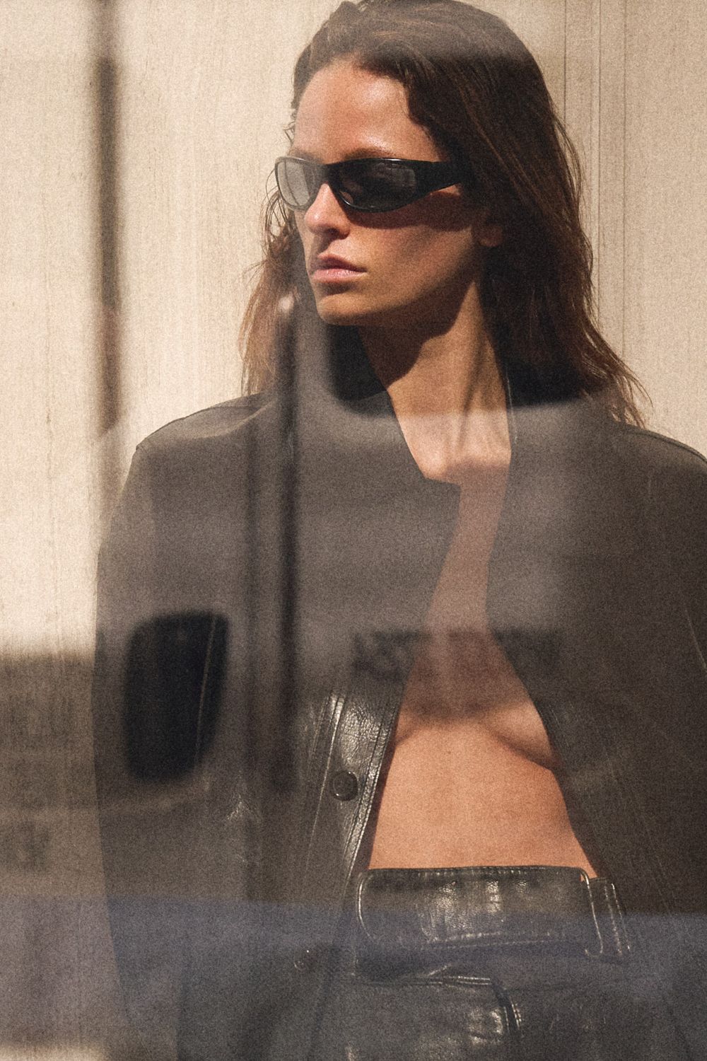 Woman in black wraparound sunglasses wearing an open leather jacket and leather trousers, photographed through a reflective window.
