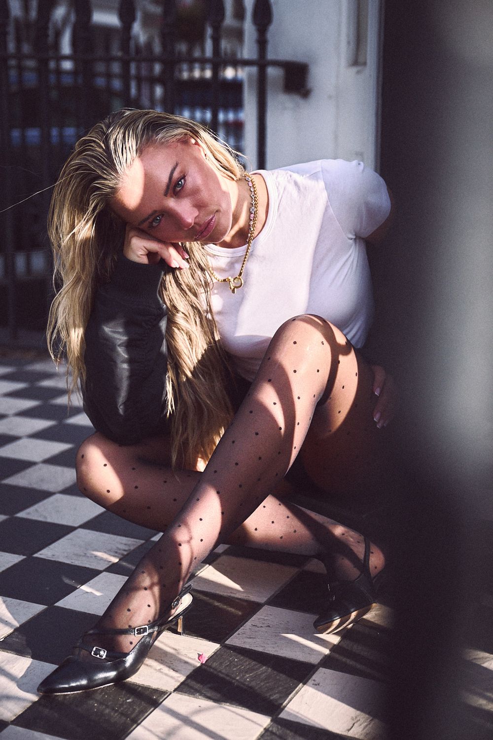 Blonde woman in a white tee, polka dot tights and black buckle shoes sits on a black and white checkered floor in warm sunlight.