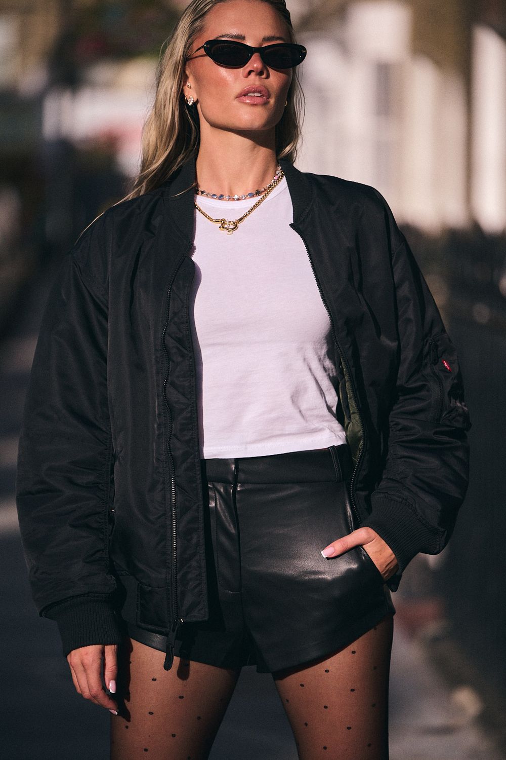 Woman in black sunglasses wears a black bomber jacket, white tee, leather shorts and polka dot tights on a city street.