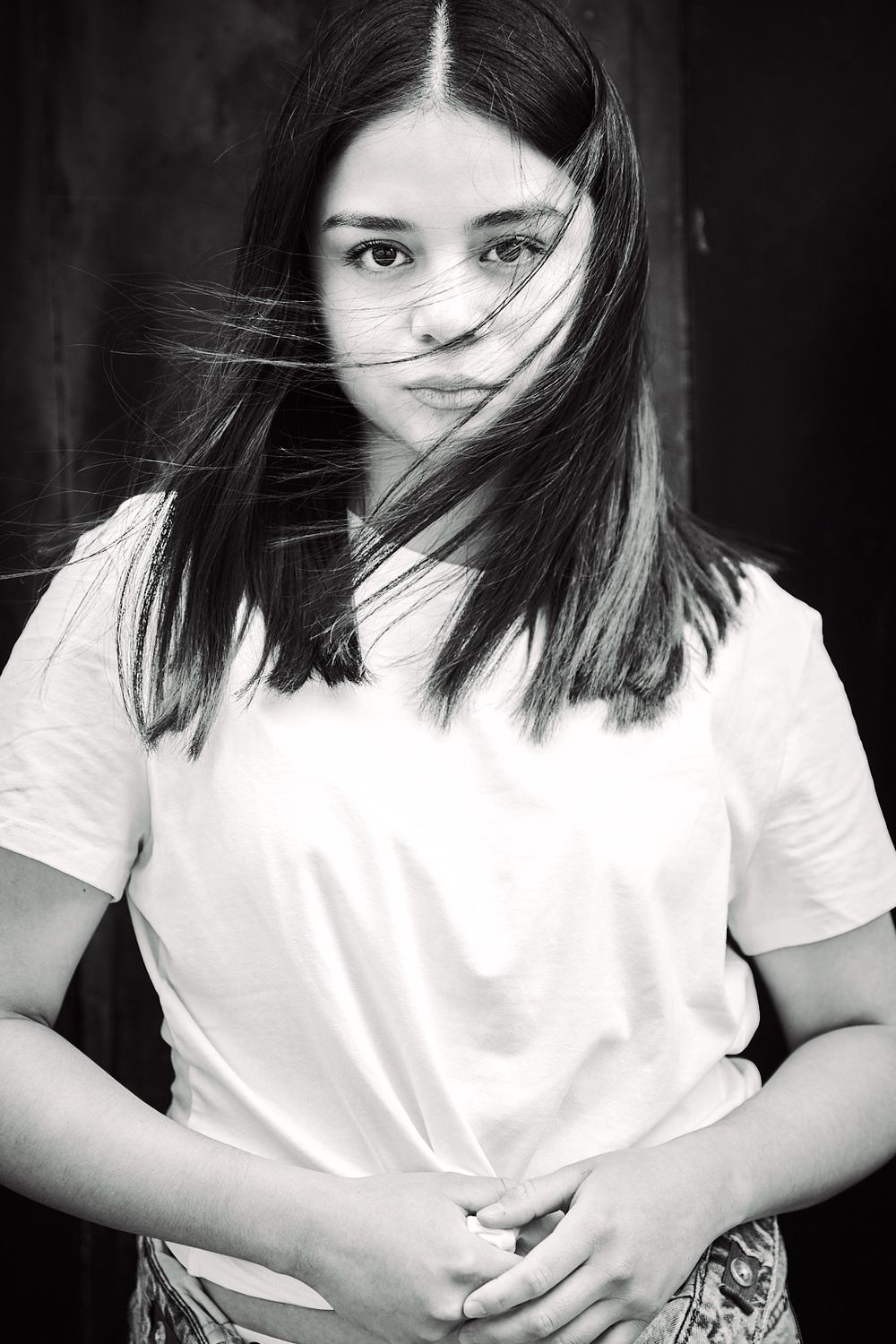 Black and white portrait of a young woman with long hair blowing across her face, wearing a white t-shirt with hands clasped in front.