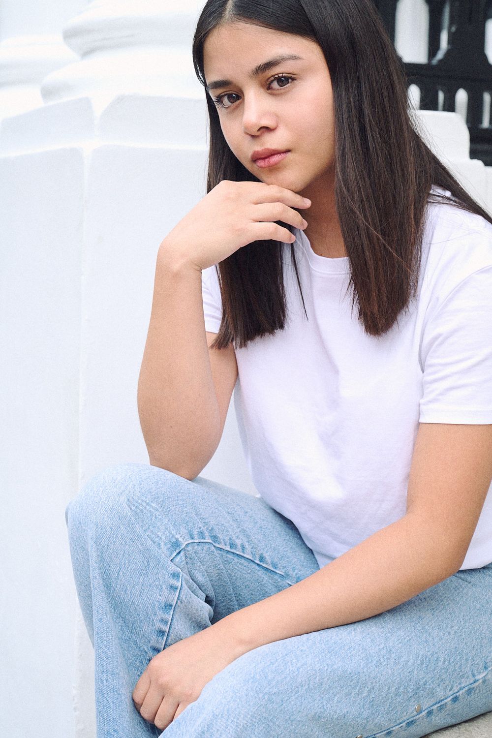 Young woman with long dark hair sits on white steps in a white t-shirt and light blue jeans, resting her hand near her chin.