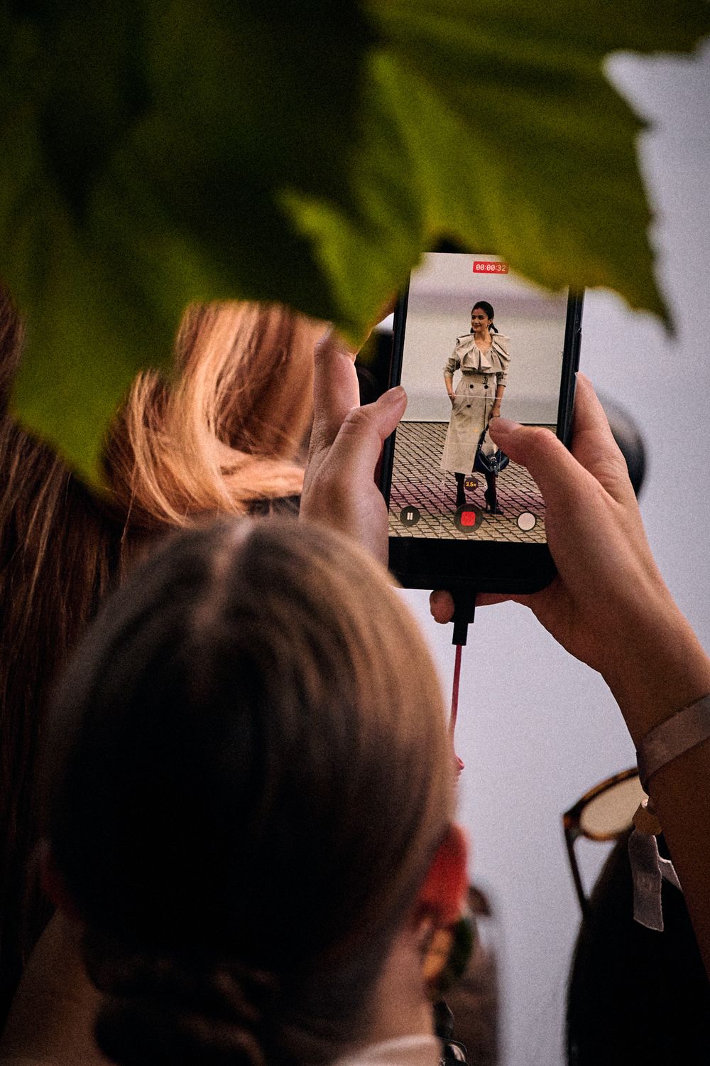 Spectators watch a fashion show through a smartphone screen showing a model in a trench coat and boots, framed by green leaves.
