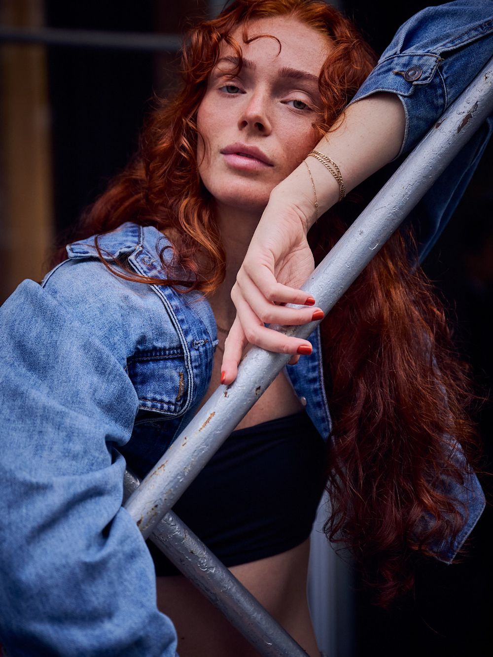 Young woman with long wavy red hair in a denim jacket and black top leans on a metal scaffolding pole, resting her chin on her hand.
