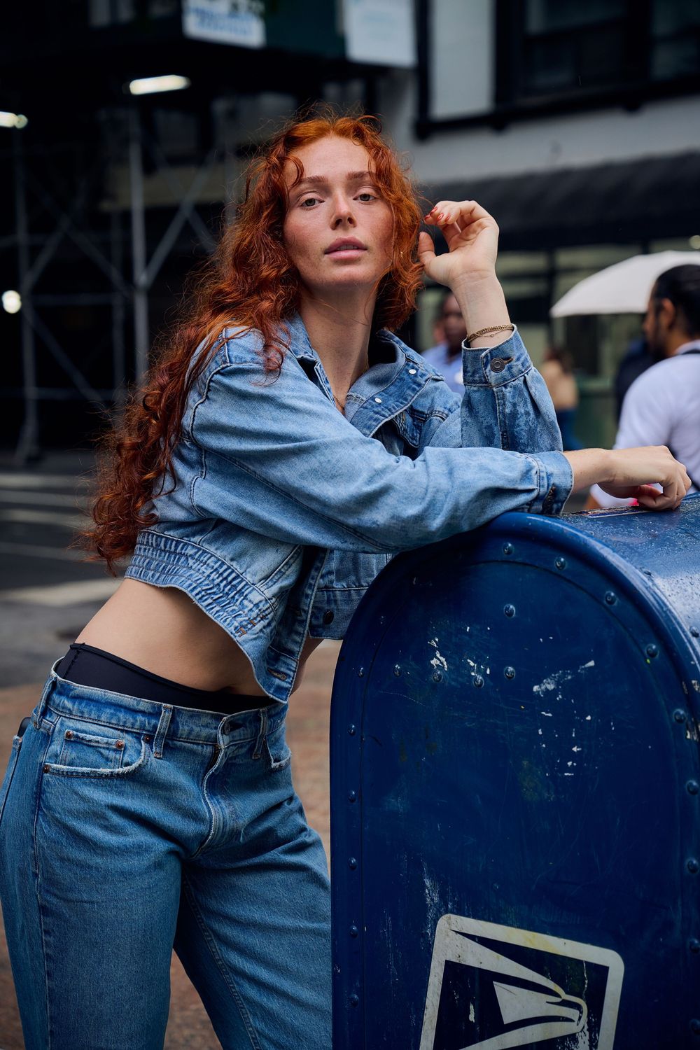 Woman with long curly red hair in a denim jacket and jeans leaning on a blue USPS mailbox on a busy city street.