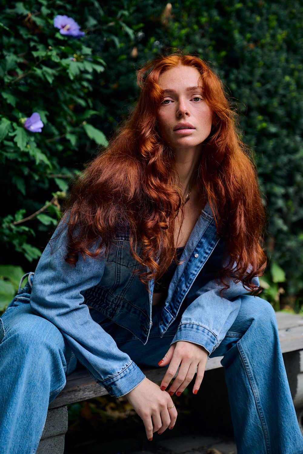 Woman with long wavy red hair in a denim jacket and jeans sitting on a wooden bench near green leaves and purple flowers.
