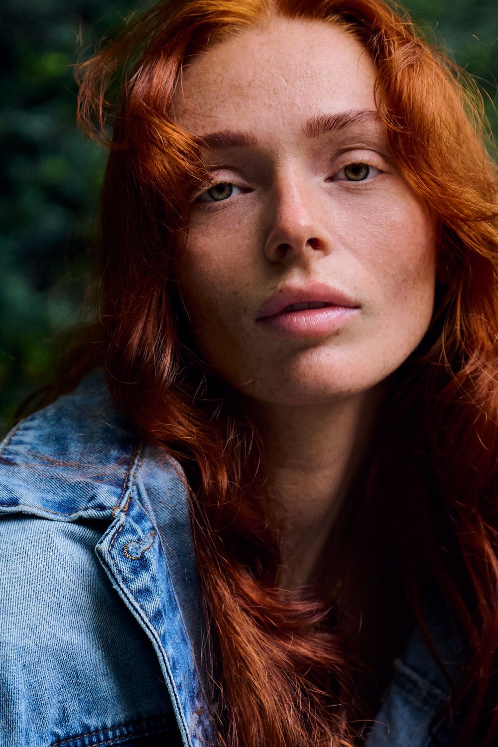 Close-up portrait of a young woman with long red hair and freckles wearing a denim jacket against a blurred green background.