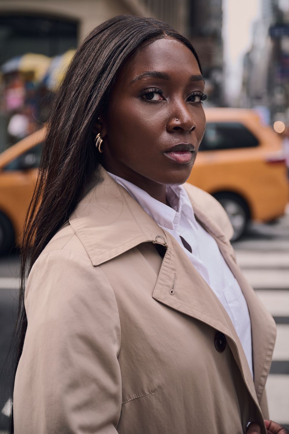 Woman with long straight hair wearing a beige trench coat and white shirt stands on a busy New York street with a yellow taxi behind her.