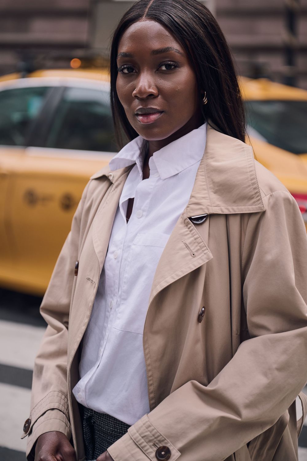 Woman in a beige trench coat and white collared shirt stands on a city street with a yellow taxi in the background.