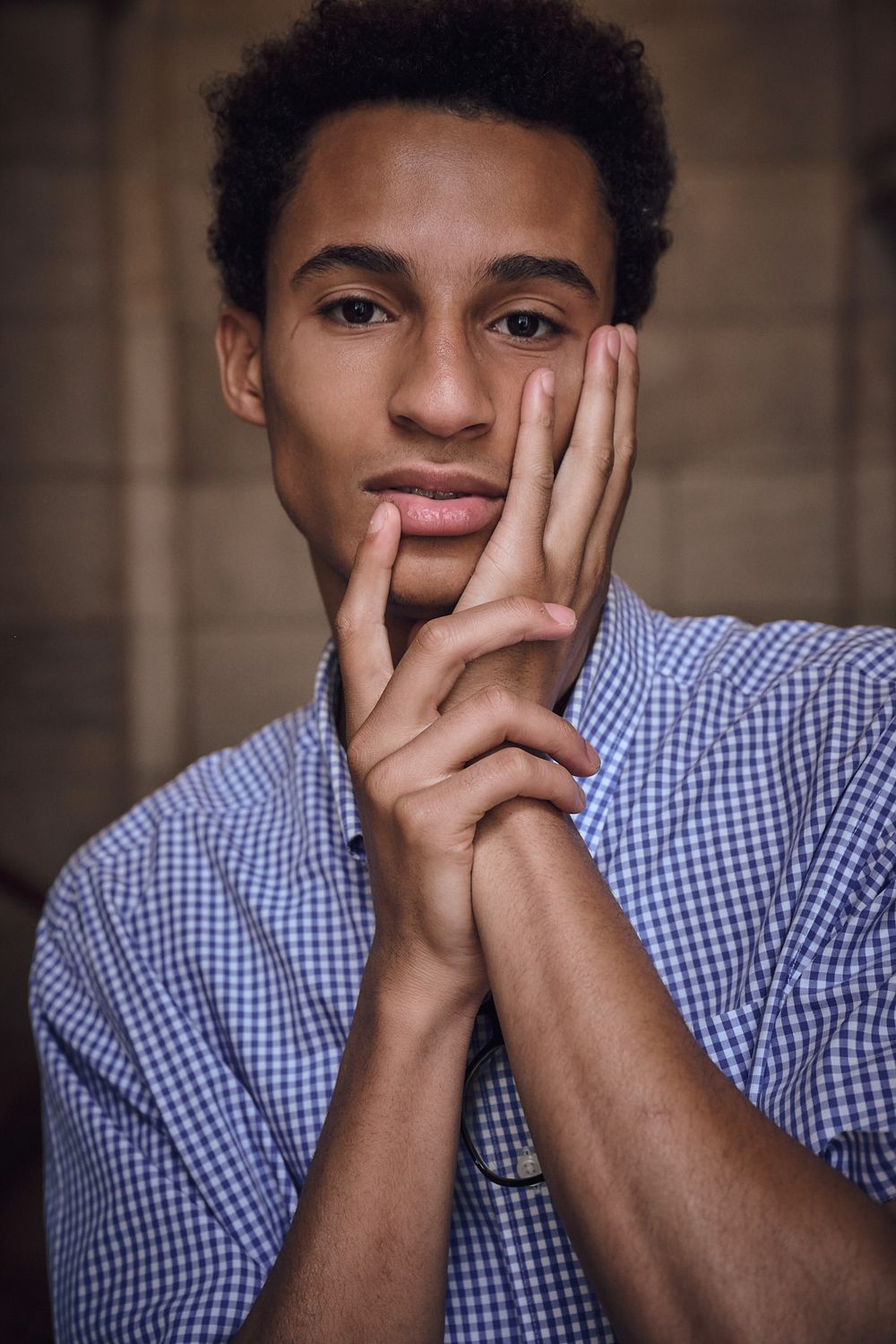 Portrait of a young man in a blue gingham shirt resting his face against his clasped hands with a calm, thoughtful expression.
