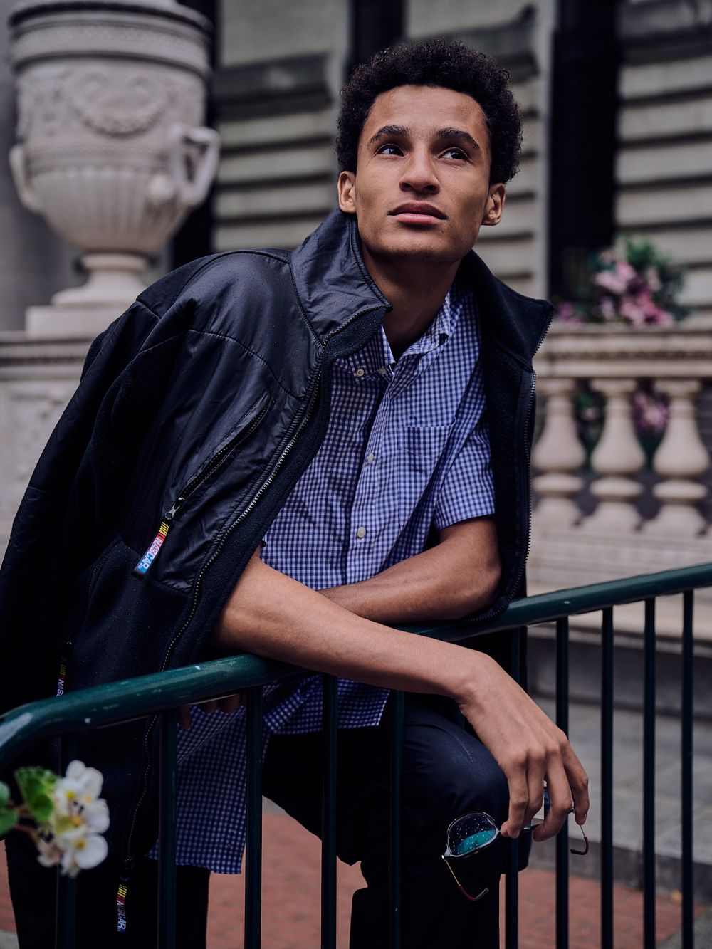 Young man in a black leather jacket and blue gingham shirt leans on a green metal railing near stone balustrades and pink flowers.
