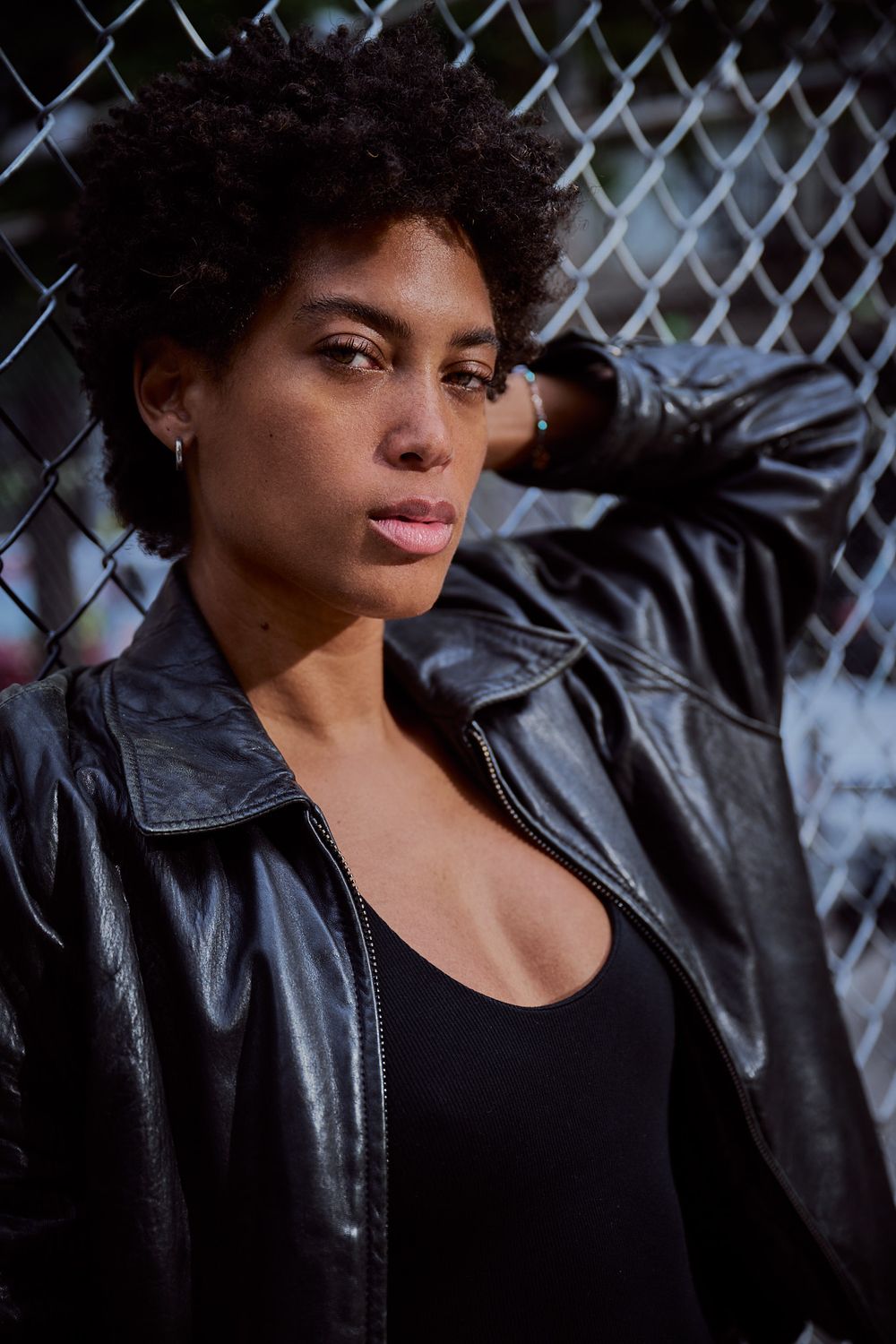 Woman with short natural curly hair wearing a black leather jacket poses with her arm raised against a chain-link fence.