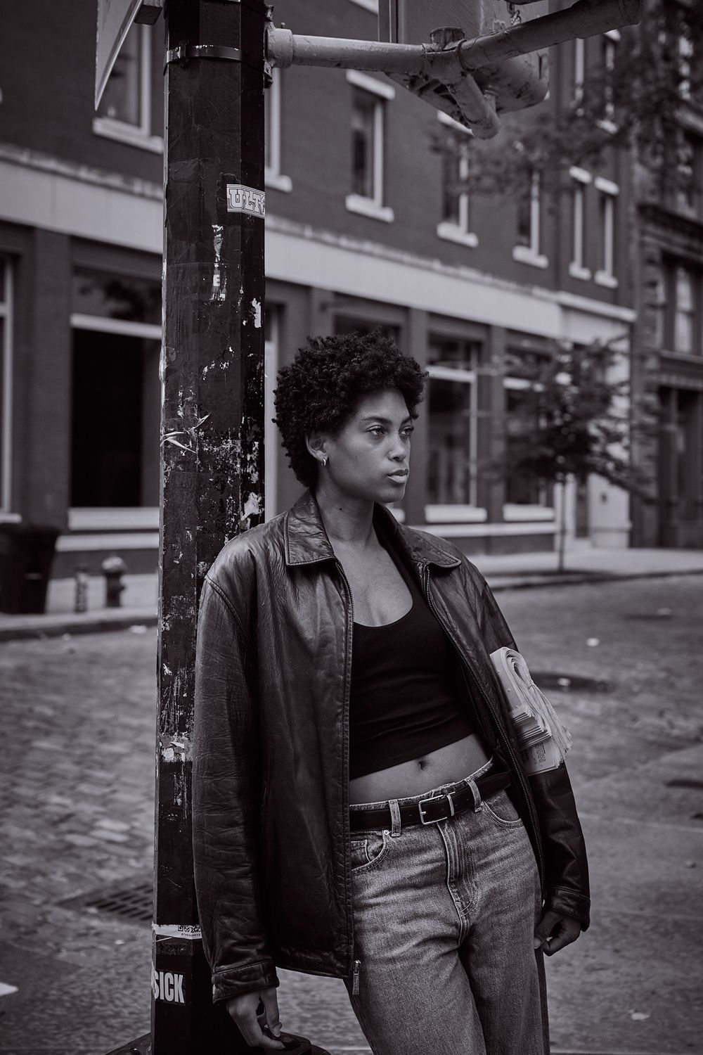 Black and white photo of a woman with short curly hair in a leather jacket and jeans leaning on a street pole on a cobblestone city street.